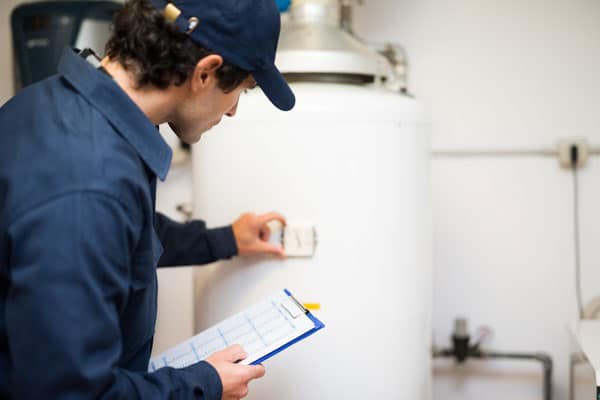 A person in a blue uniform and cap inspects a broken water heater, adjusting a control and holding a clipboard with a checklist in a utility room.