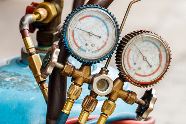 Two pressure gauges with colored rims, attached to a blue metal tank and brass valves, display readings for propane home heating. Various hoses and fittings are connected to the equipment.