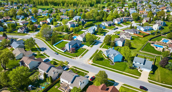 Aerial view of a suburban neighborhood with single-family homes, green lawns, tree-lined streets, and cars parked in driveways and along the roads. The area looks orderly and well-maintained.