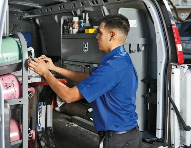 A man in a blue shirt stands next to an open van, working with tools and equipment stored inside. Various tanks, containers, and toolboxes are organized along the van’s shelving.