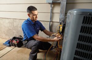 A technician in a blue shirt kneels next to an outdoor HVAC unit, using tools to check wires. A tool bag with equipment is on the ground beside him, and the unit is installed next to a building wall.