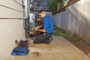 A technician in a blue shirt kneels on a paved walkway next to a building, servicing an outdoor HVAC unit with tools and equipment laid out beside him.