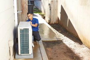 A technician in a blue shirt kneels on a concrete path beside a house, inspecting or repairing an outdoor air conditioning unit.