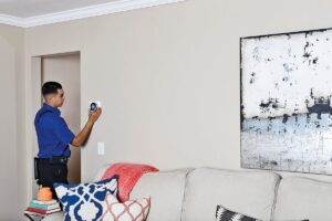 A person in a blue shirt stands near a doorway, installing or inspecting a wall-mounted device in a living room with a beige sofa, patterned pillows, and an abstract painting on the wall.
