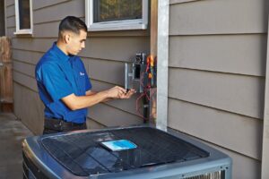 A man in a blue shirt uses tools to work on an electrical panel mounted on the exterior wall of a house, next to an outdoor air conditioning unit.