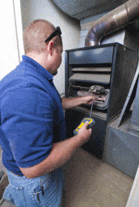 A person wearing a blue shirt and safety glasses uses a handheld device while inspecting the inside of a furnace unit, with tools and metal ductwork visible around the equipment.
