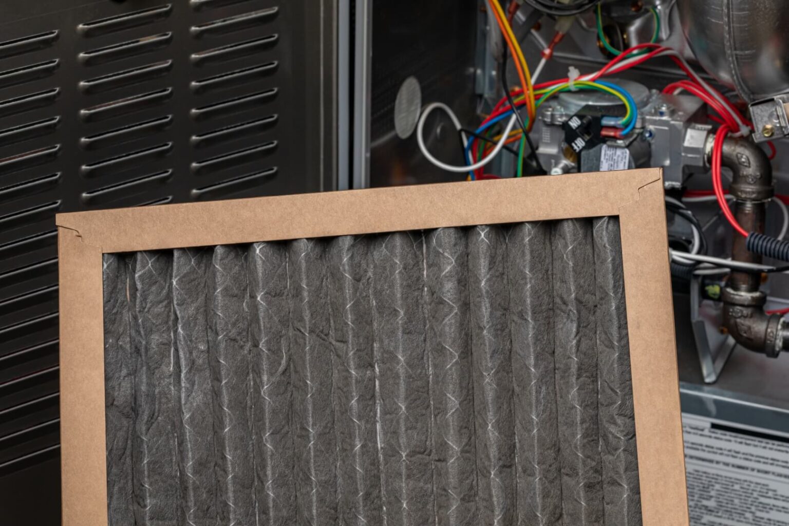 A dirty, used air filter is held in front of an open furnace panel showing wires and internal components—a clear sign it’s time for Air Conditioning Maintenance San Diego homeowners can trust. The filter appears dark with dust and debris.