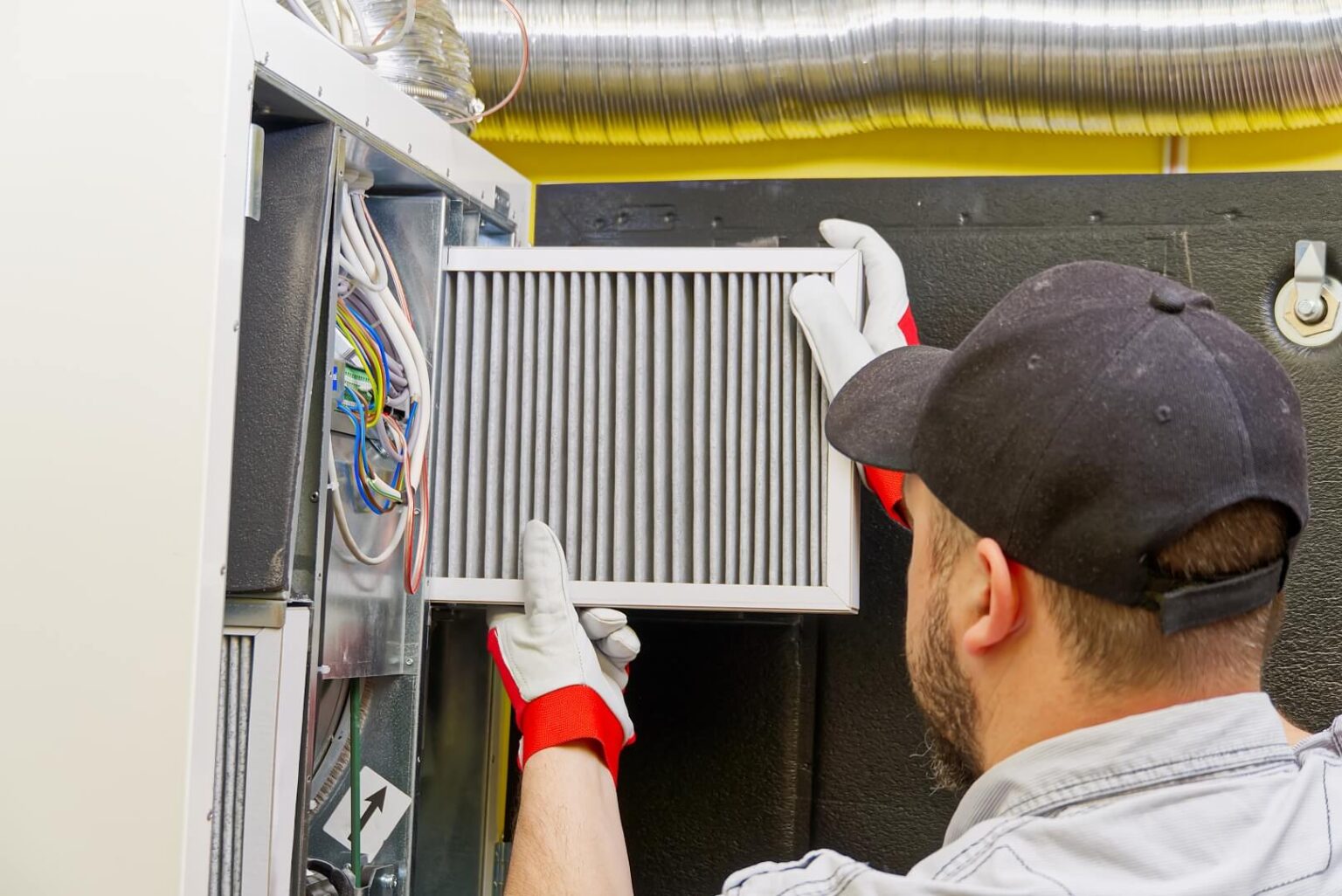 A person wearing gloves and a cap is inserting a clean air filter into an HVAC unit during Air Conditioning Maintenance San Diego, with visible wires and ductwork in the background.