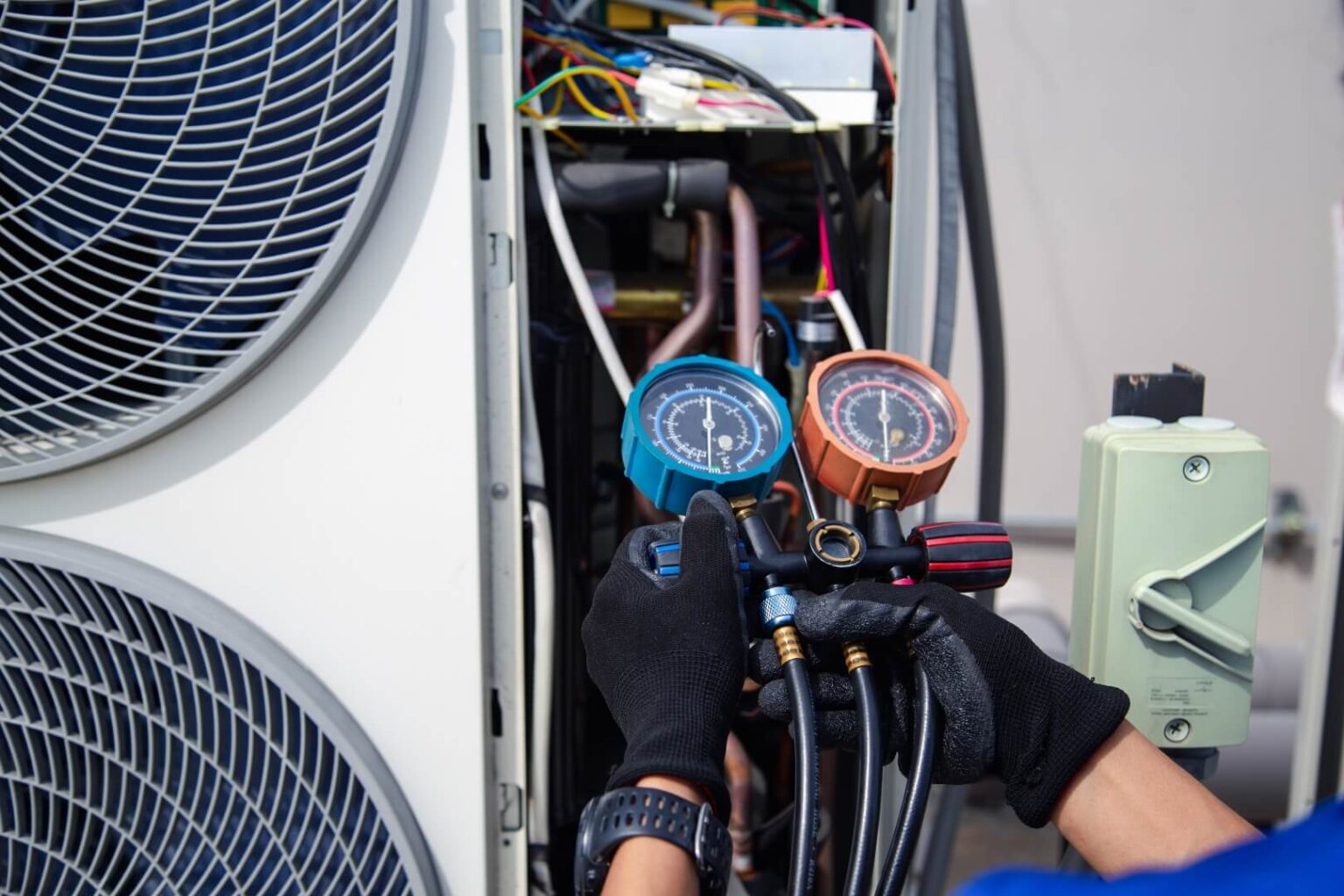 A technician wearing black gloves uses a manifold gauge to check refrigerant levels on an outdoor HVAC unit during an Air Conditioning Repair Poway service. The unit’s internal wiring and large cooling fans on the left are clearly visible.