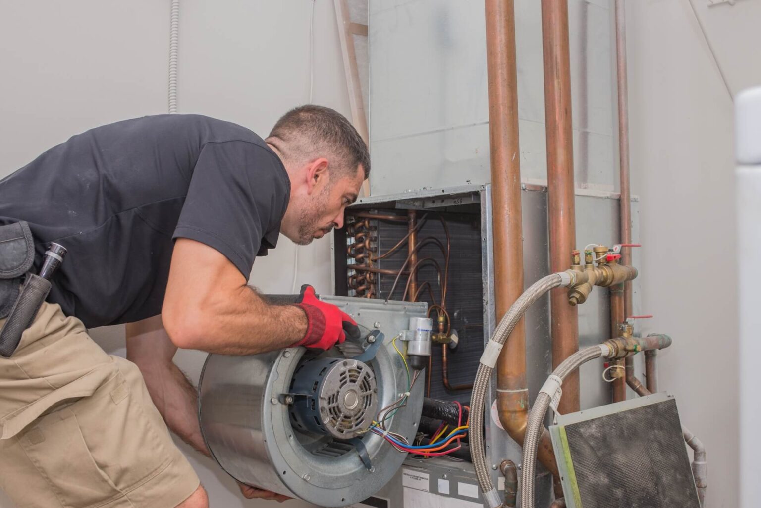 A technician in red gloves and a black shirt is performing Air Conditioning Repair in Serra Mesa, servicing the blower assembly of an HVAC unit with copper pipes and wiring visible in the background.