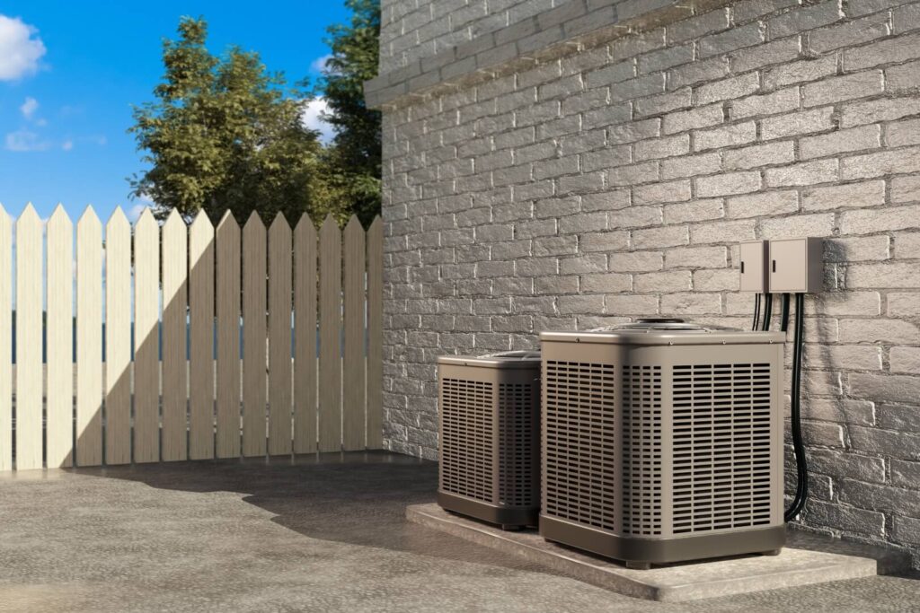 Two outdoor HVAC units, part of our expert cooling services, are set on a concrete pad beside a gray brick wall. Electrical wiring runs above, while a wooden picket fence and trees appear in the background under the blue sky.