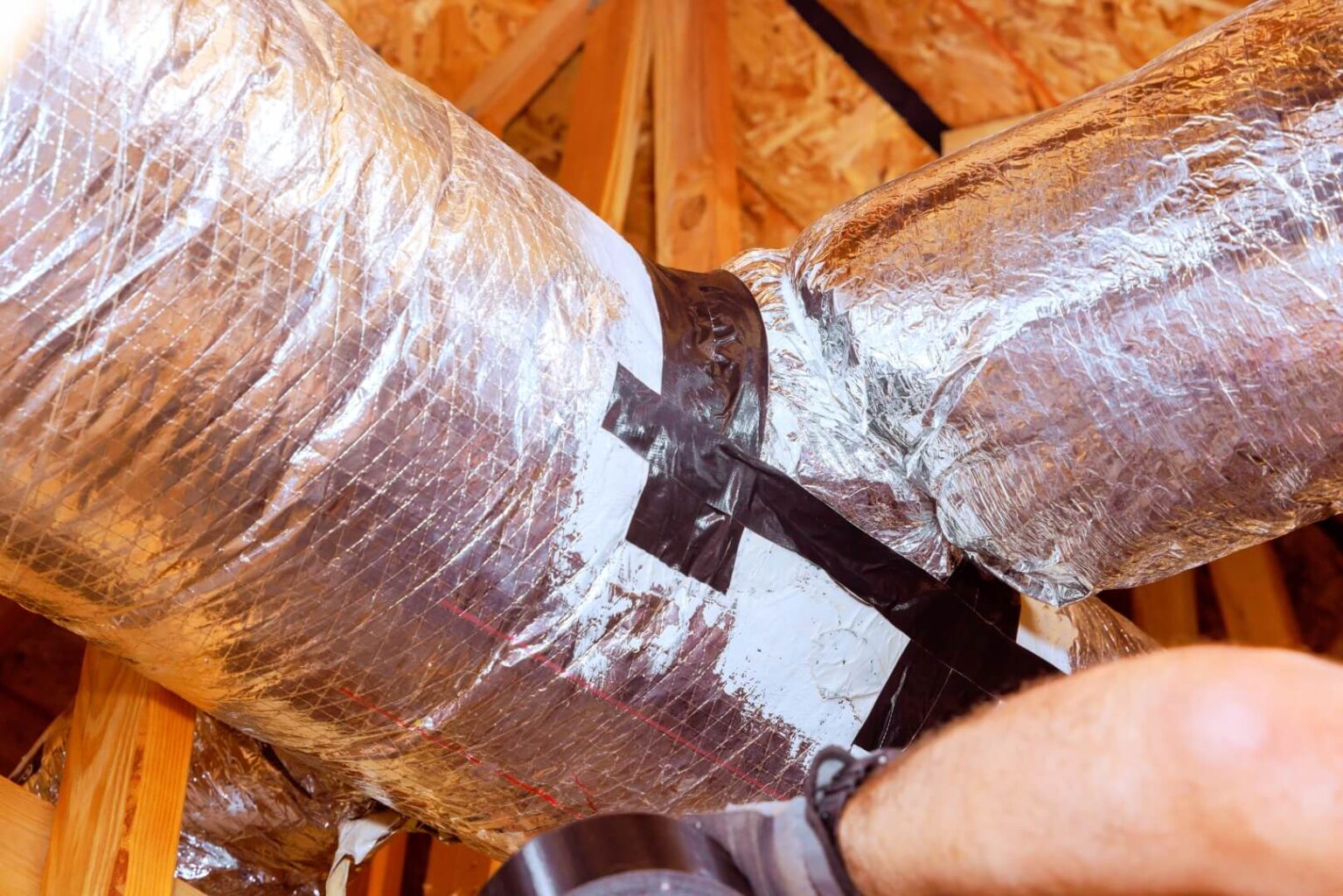 Close-up of HVAC ductwork in an attic, wrapped in reflective insulation and sealed with black duct tape at the joints, highlighting air duct sealing, with part of a person’s arm visible in the lower right corner.