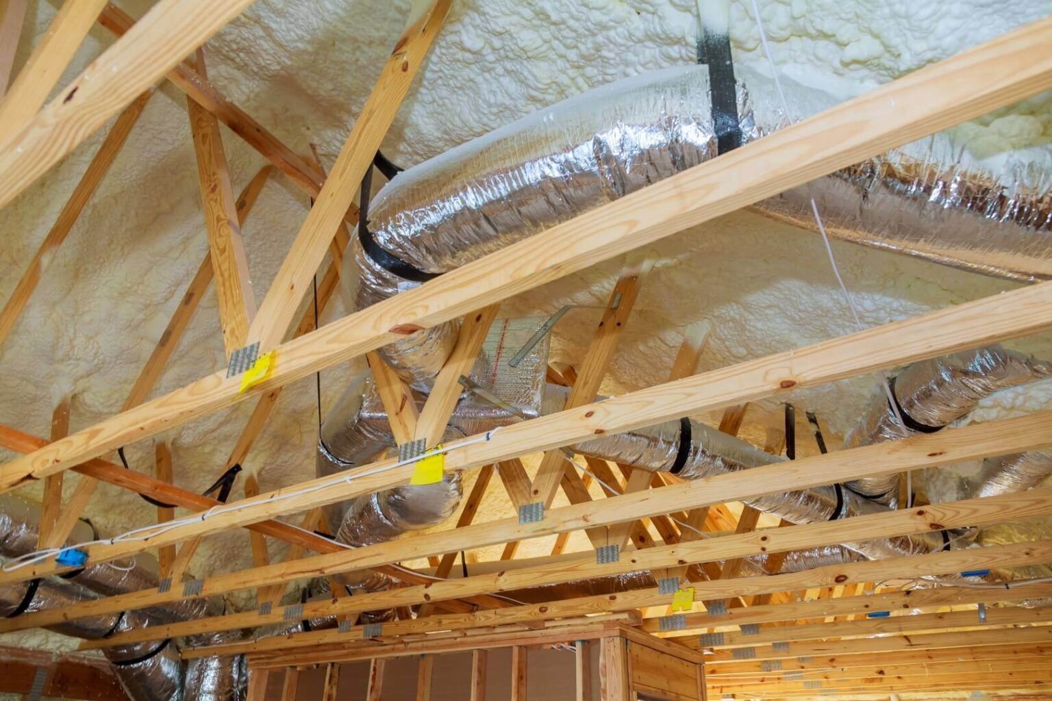 Exposed wooden roof trusses with shiny metal HVAC ductwork and spray foam insulation highlight the importance of proper HVAC sealing in a building under construction.