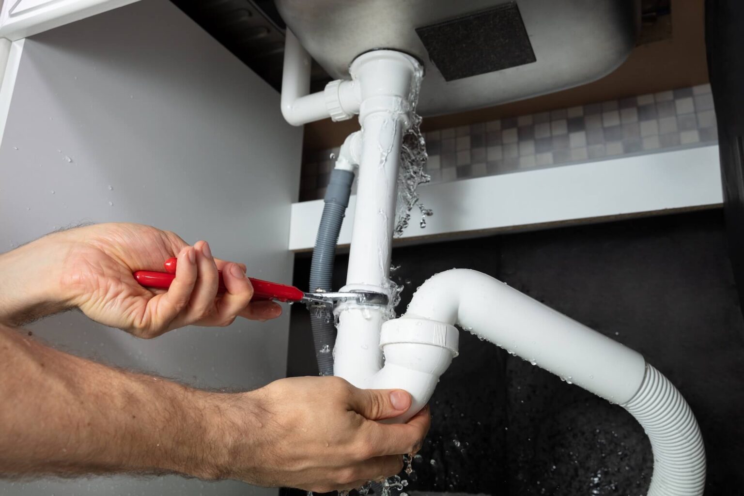 A person uses a red wrench to tighten a leaking pipe under a sink, working quickly on burst pipe repair as water sprays from the connection.