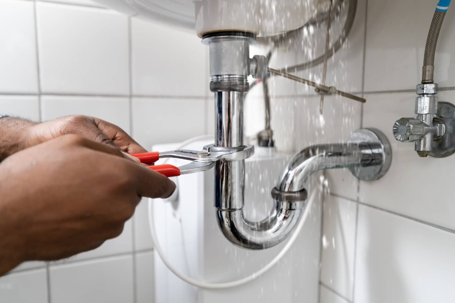 A person uses pliers to fix a burst pipe under a sink. Water sprays from the connection during the burst pipe repair, with white tiled walls and additional plumbing visible in the background.