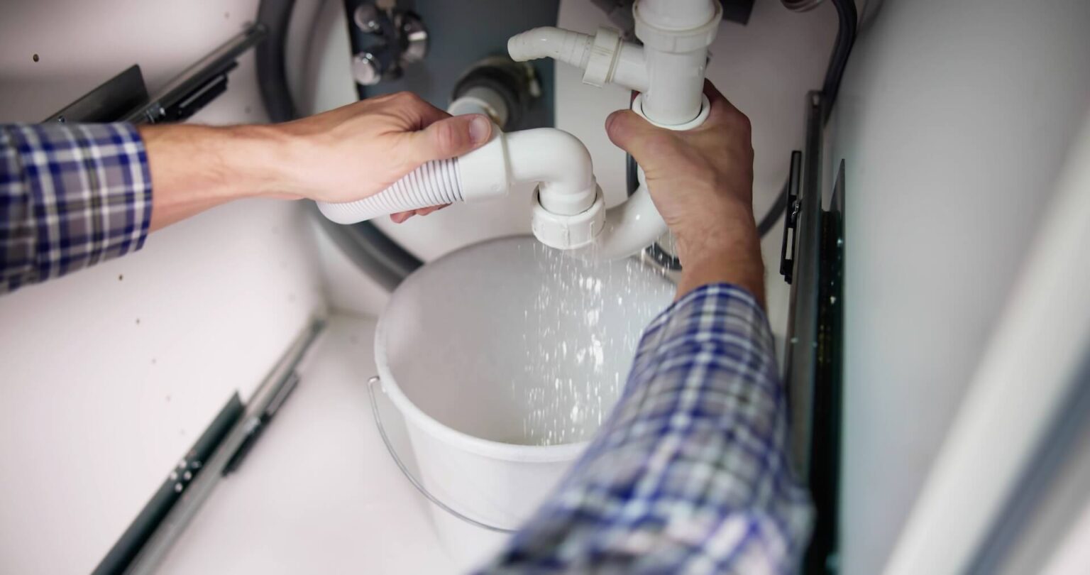 A person in a plaid shirt is fixing plumbing under a sink, performing burst pipe repair as they hold a white drain pipe over a bucket while water pours out.