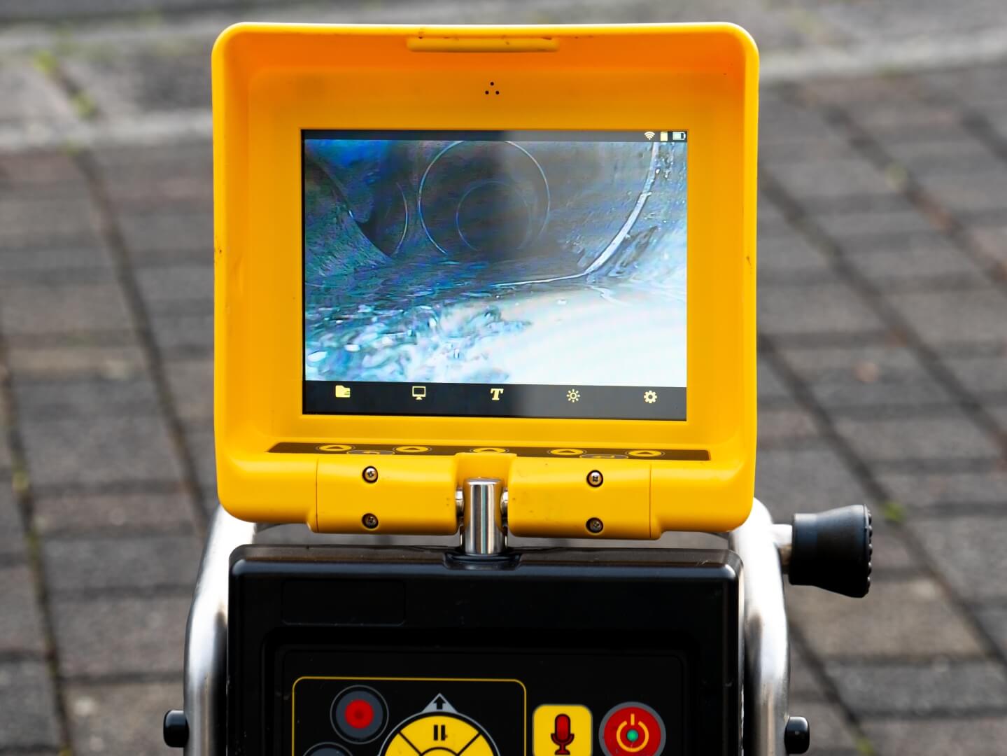 A yellow inspection camera monitor displays a live video feed of the inside of a pipe. The device, used by a Camera Inspection Service San Diego, is set up outdoors on a paved surface with various control buttons below the screen.