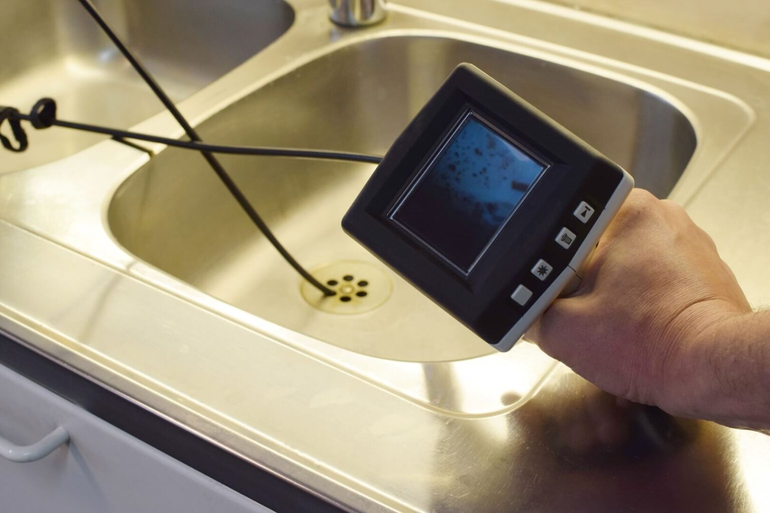 A person uses an inspection camera with a flexible cable to examine the inside of a stainless steel sink drain, demonstrating the thorough approach of a Camera Inspection Service San Diego offers.