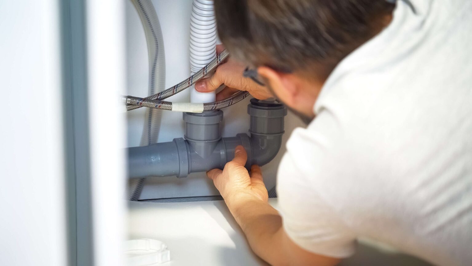 A person in a white shirt is installing or repairing grey plumbing pipes under a sink, possibly performing drain cleaning in Bay Park, CA, using both hands to adjust the connections. Hoses and pipes are visible in the background.
