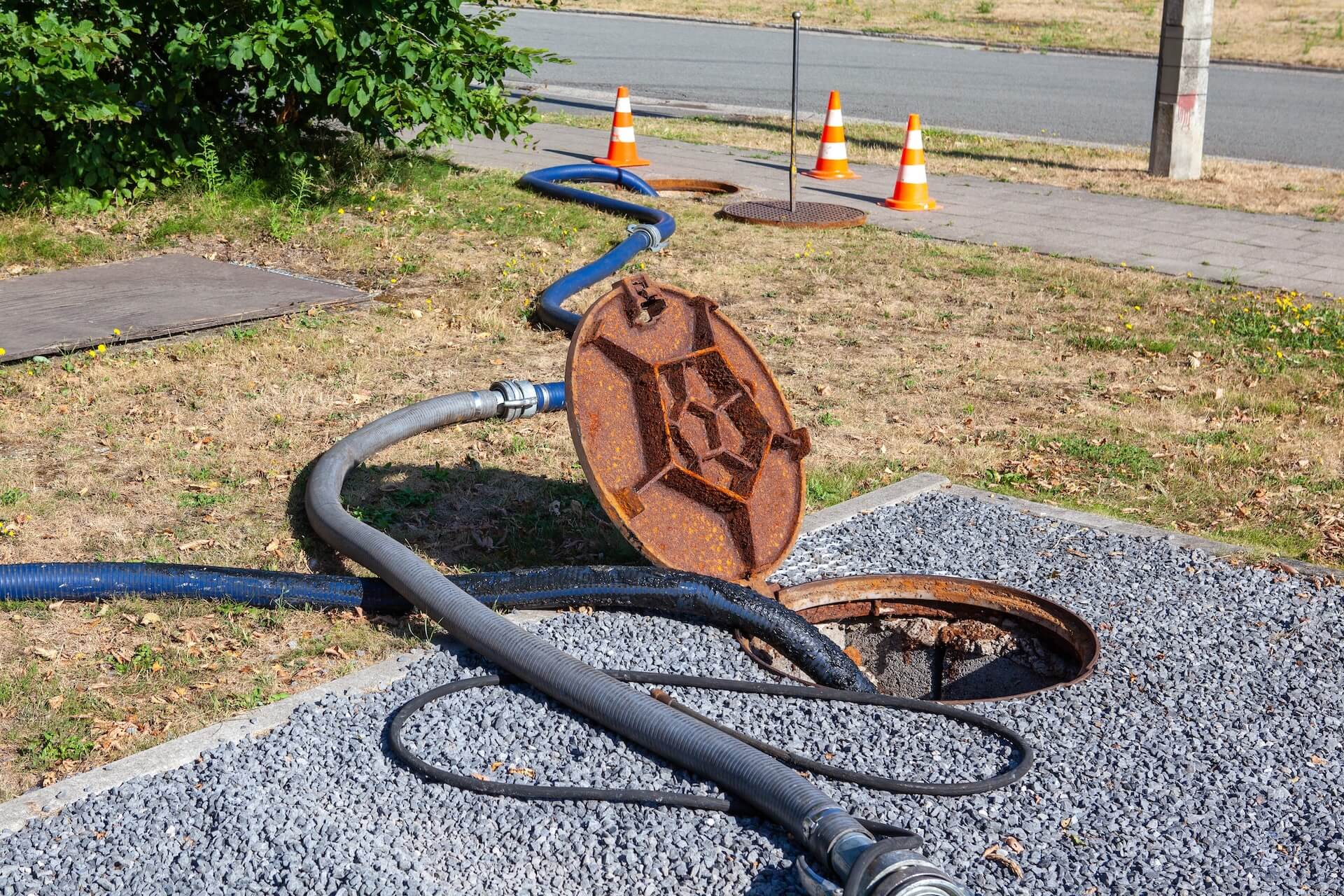 A rusty manhole cover is propped open with hoses running into the opening. Orange traffic cones are set up on the sidewalk and grass nearby, marking a Drain Clearing San Diego maintenance scene next to the road.