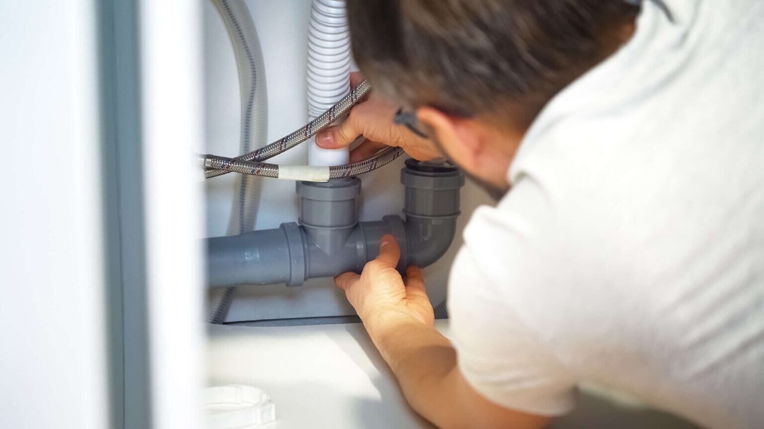 A person wearing glasses works under a sink, inspecting or tightening plumbing pipes and fittings with both hands. Flexible hoses and plastic pipes are visible, suggesting expert drain cleaning Escondido services in action.