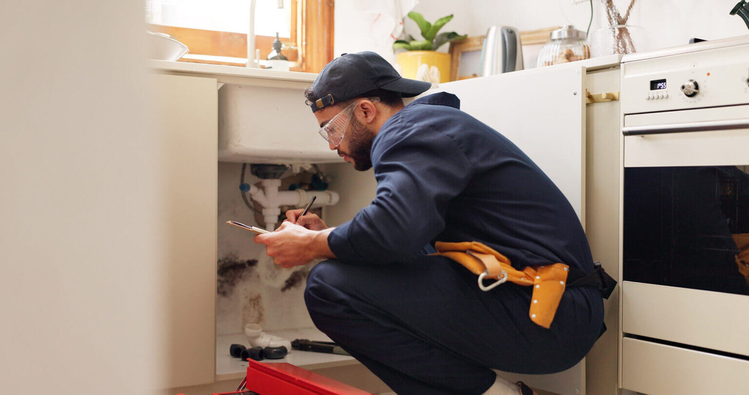 A person wearing a navy jumpsuit, cap, and safety glasses is kneeling and writing on a clipboard while inspecting plumbing under a kitchen sink—tools and drain cleaning Fallbrook supplies are visible nearby.