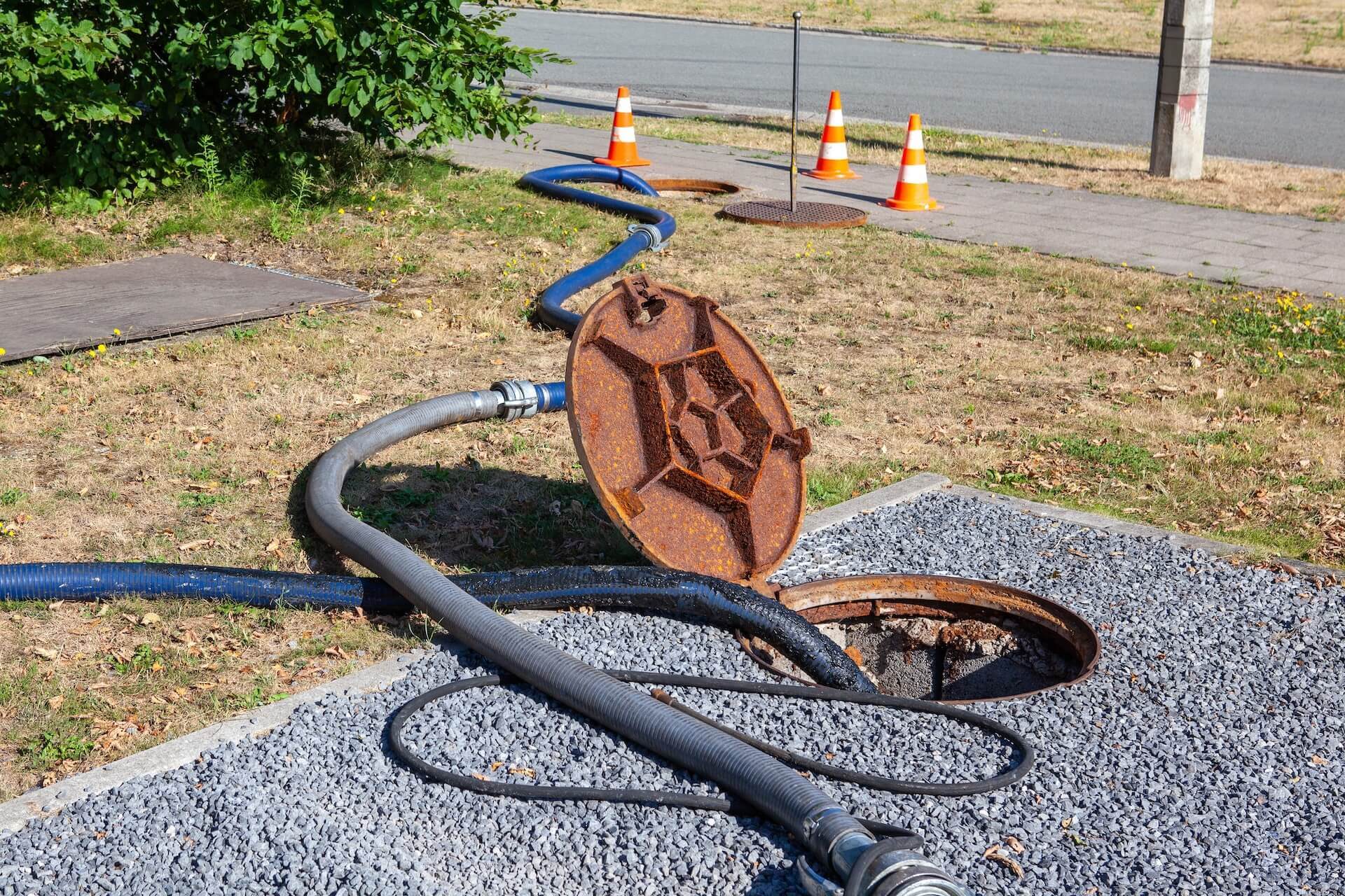 A manhole cover is open with large hoses running into it for drain cleaning La Jolla services. Traffic cones are set up nearby on the sidewalk, indicating ongoing maintenance work. The area around the manhole is covered with gravel and grass.