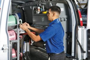 A man in a blue uniform works inside the open back of a service van, adjusting equipment and supplies organized on shelves. Various tools and canisters are visible around him.