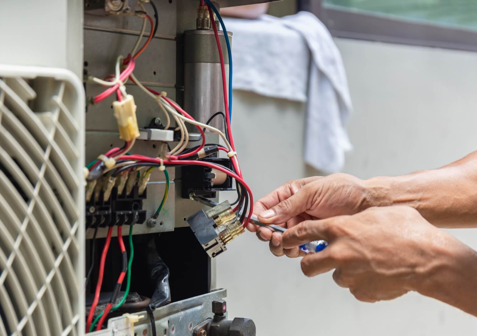 A person uses both hands to repair or connect electrical wires inside an open air conditioning unit, with various colored wires and components visible—a common scene during furnace repair or heating repair tasks.