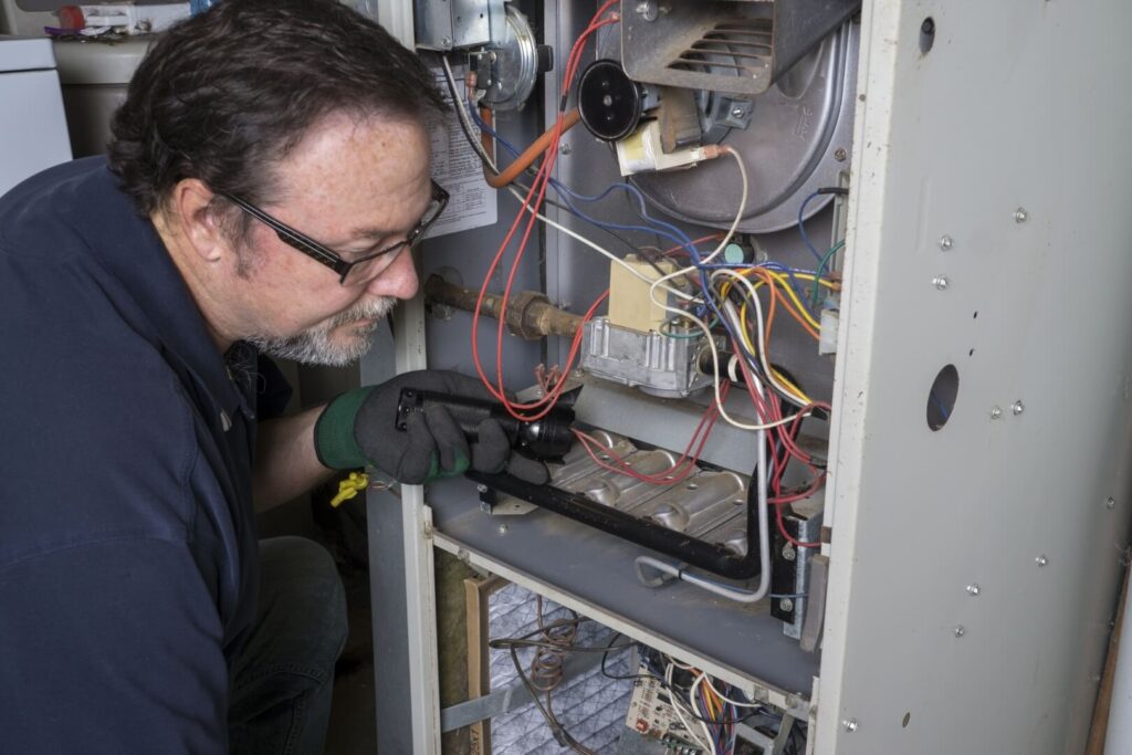 A man wearing glasses and gloves uses a flashlight to inspect the wiring and components inside an open furnace or HVAC unit during routine furnace maintenance San Diego homeowners trust.