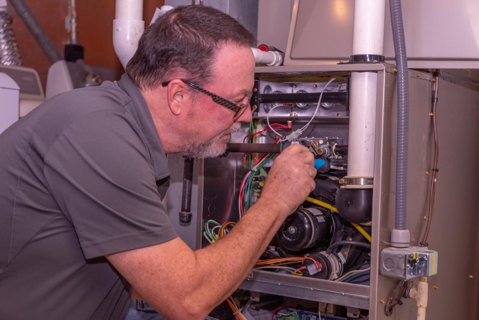 A man wearing glasses and a gray shirt uses a tool to repair or inspect an open furnace or HVAC unit, exposing internal components and wiring—perfectly capturing furnace maintenance San Diego professionals provide.
