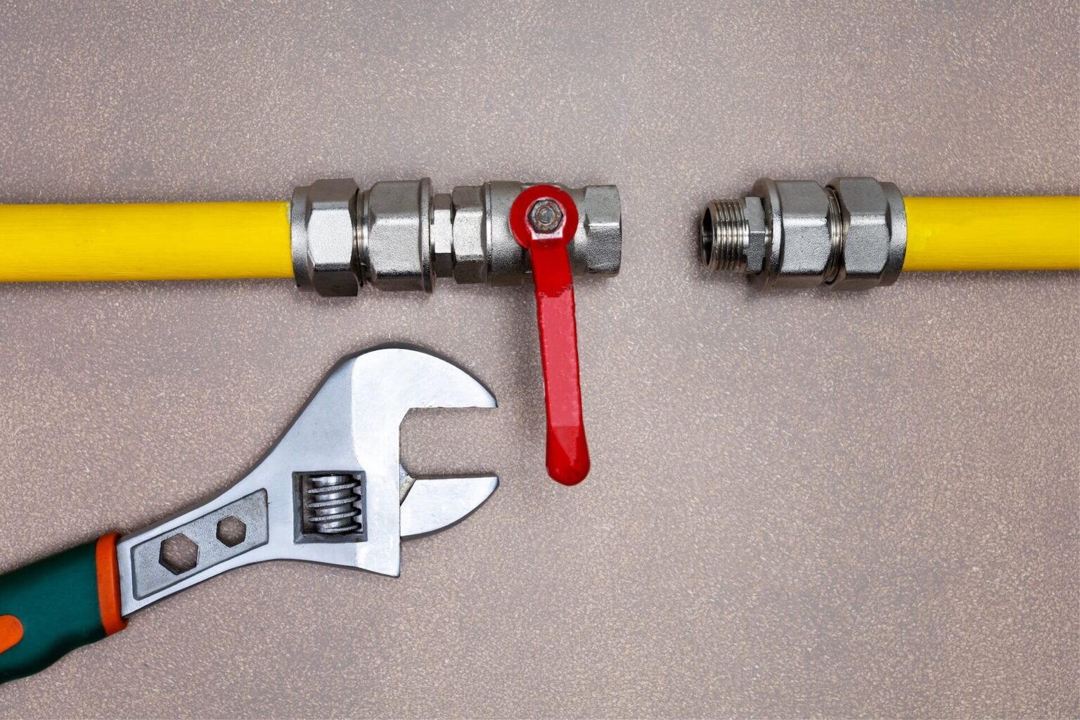 A yellow gas pipe with a red valve handle is disconnected, showing threaded ends—ideal for Gas Line Install San Diego needs. An adjustable wrench lies below on a brown surface.