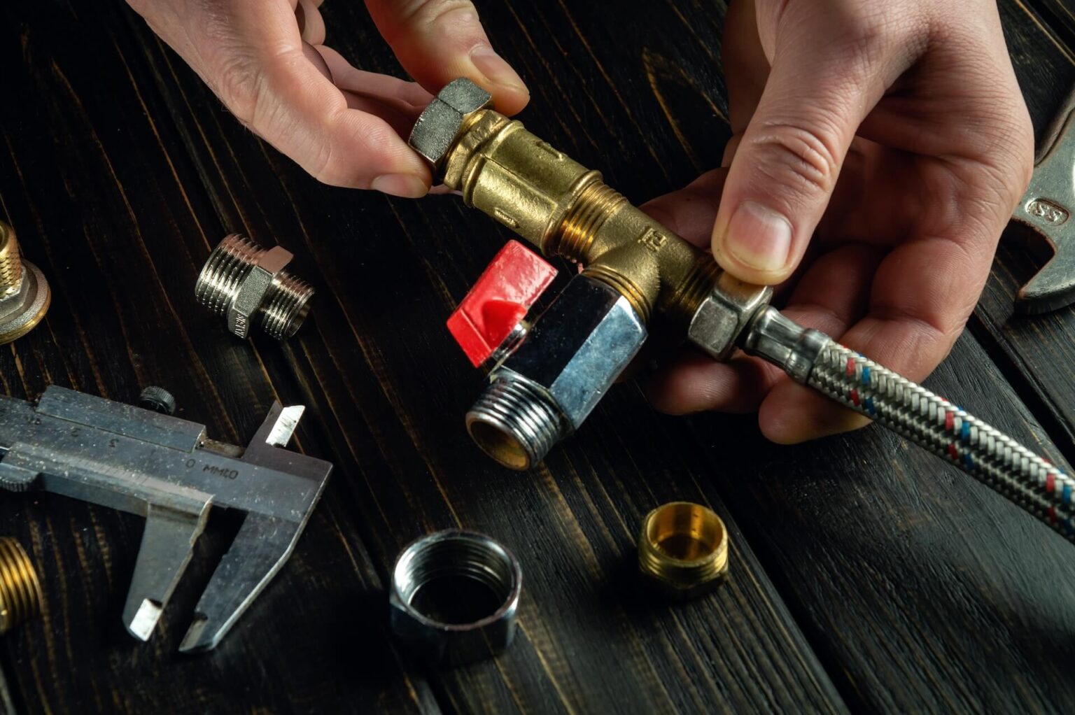 A person’s hands assemble plumbing fittings, including a brass T-joint and a red-handled valve, on a dark wooden surface with tools and metal connectors scattered nearby.