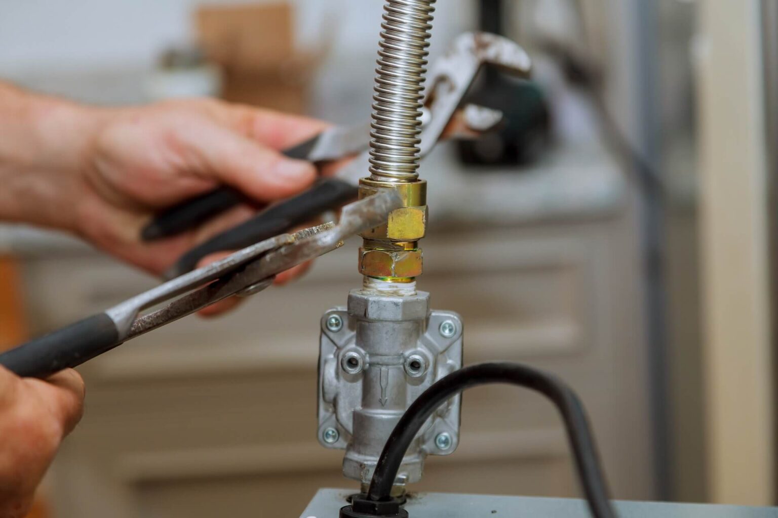 A person uses two wrenches to tighten a metal gas fitting, which is connected to a flexible hose and a valve. The background shows an out-of-focus indoor setting.