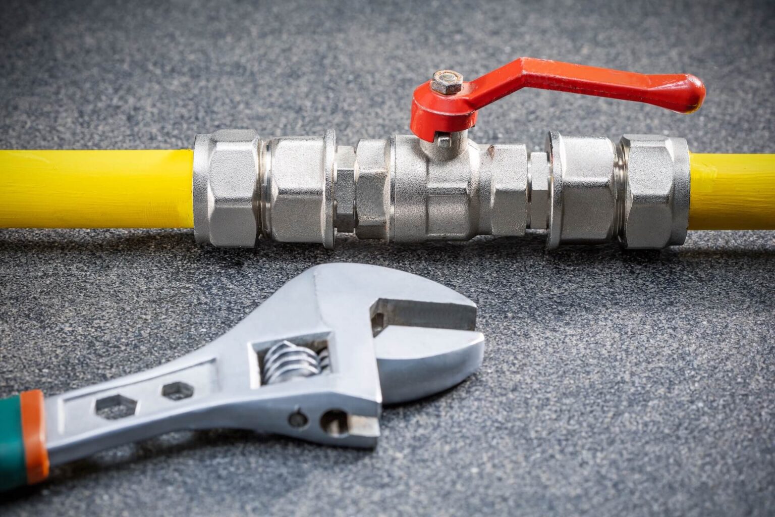 A metal valve with a red handle is connected to a yellow pipe on a gray surface. An adjustable wrench lies below the valve, positioned horizontally.