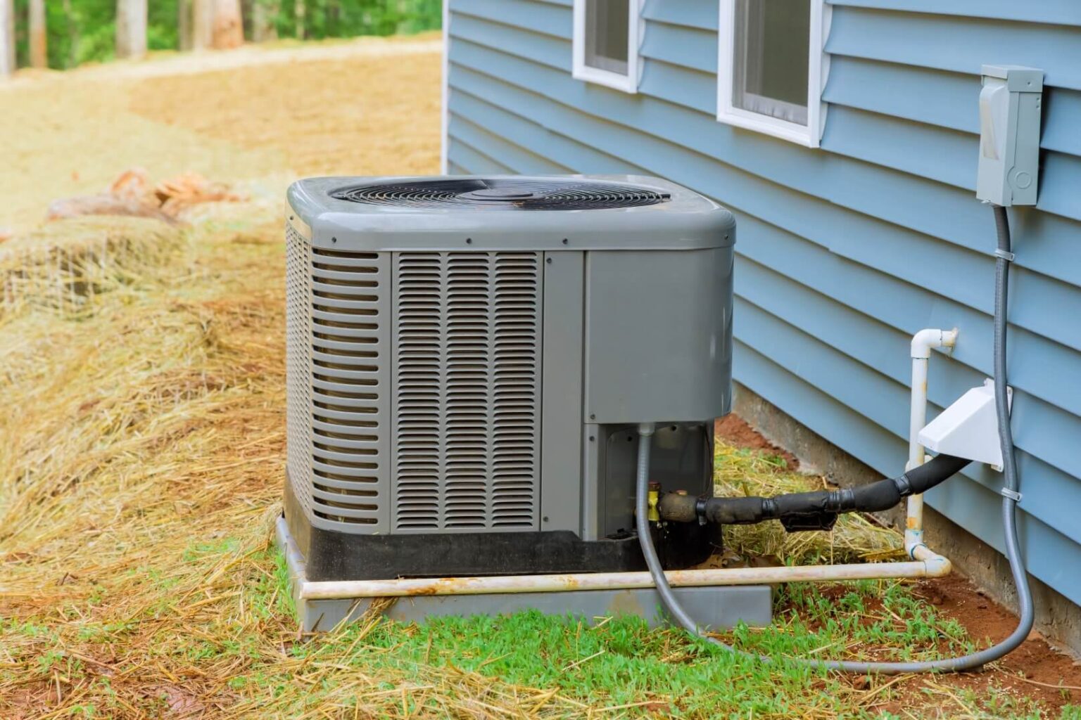 An outdoor air conditioning unit sits on a concrete slab next to a blue house with vinyl siding, pipes and cables connected—ideal for expert Heat Pump Repair San Diego services—with some dry grass covering the ground nearby.