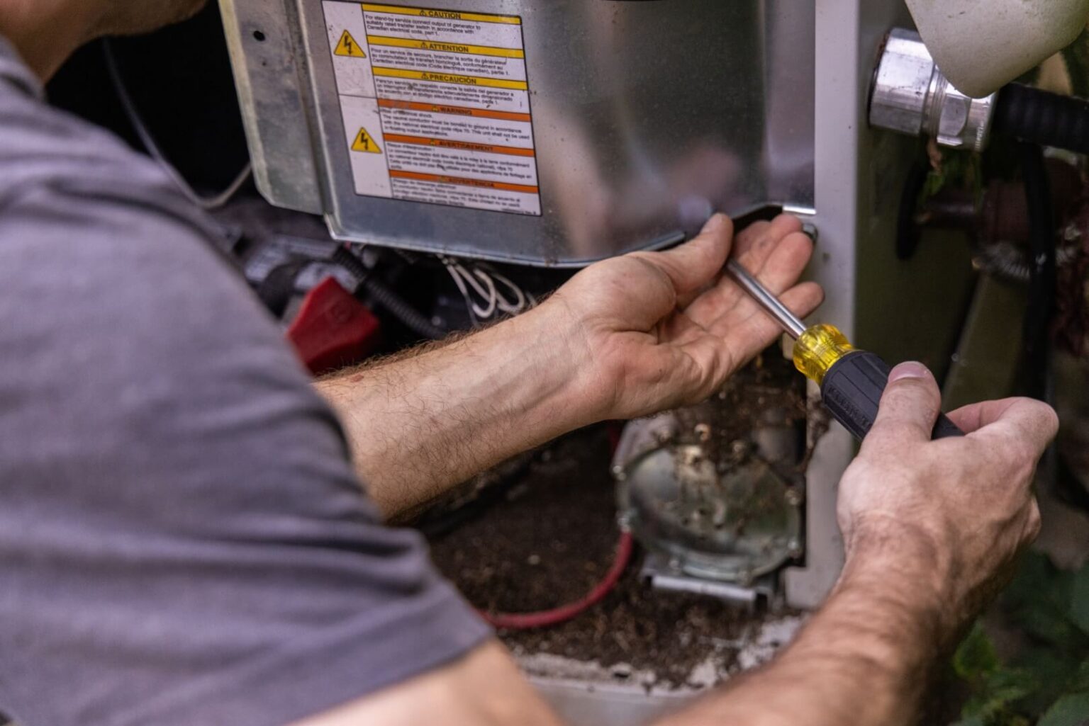 A person uses a screwdriver to remove a screw from the metal panel of a heating repair Fallbrook machine, with warning labels visible and wires exposed inside the equipment.
