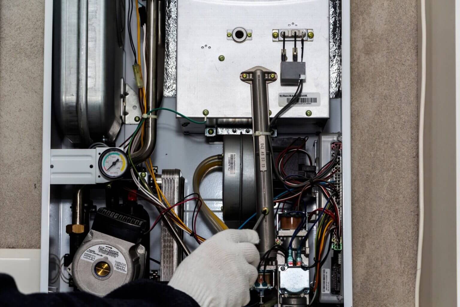 A person wearing a white glove adjusts internal wiring and components of an open gas boiler, showing wires, circuit boards, pipes, and a pressure gauge—an example of expert heating repair in North Park.