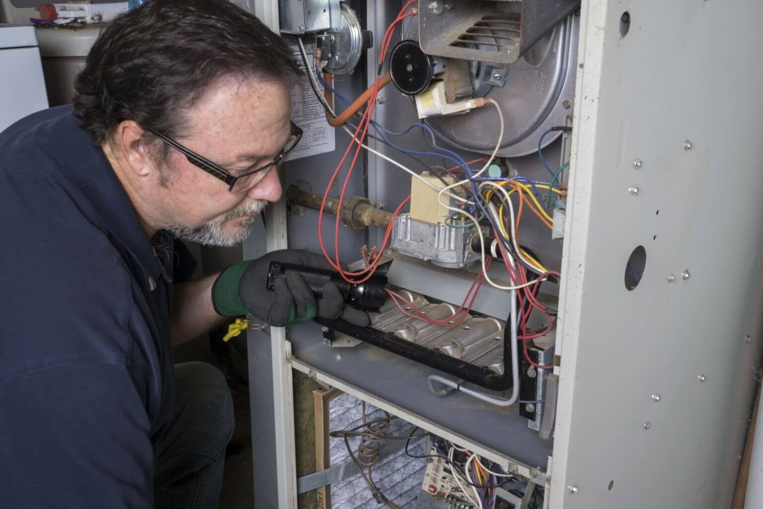 A man wearing glasses and gloves uses a flashlight to inspect the wiring and components inside a furnace or HVAC system, performing a detailed heating repair Vista service. Various colored wires and metal parts are visible inside the open unit.