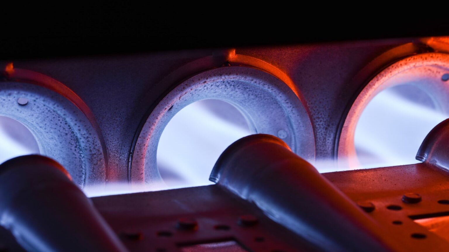 Close-up view of three gas burner tubes inside a furnace, with blue flames visible and an orange glow around the openings, showcasing expert Heating Services San Diego and the effects of intense heat on the metal surface.