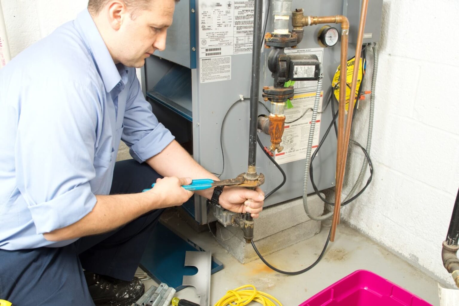 A person in a blue shirt uses a wrench to adjust a pipe connected to a furnace in a utility room, showcasing expert Heating Services San Diego. Tools and cables are visible on the floor nearby.