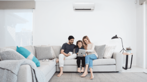 A man, woman, and child sit together on a light gray sectional sofa in a bright living room, looking at a tablet. The room features a floor lamp, blankets, and a wall-mounted air conditioning unit.