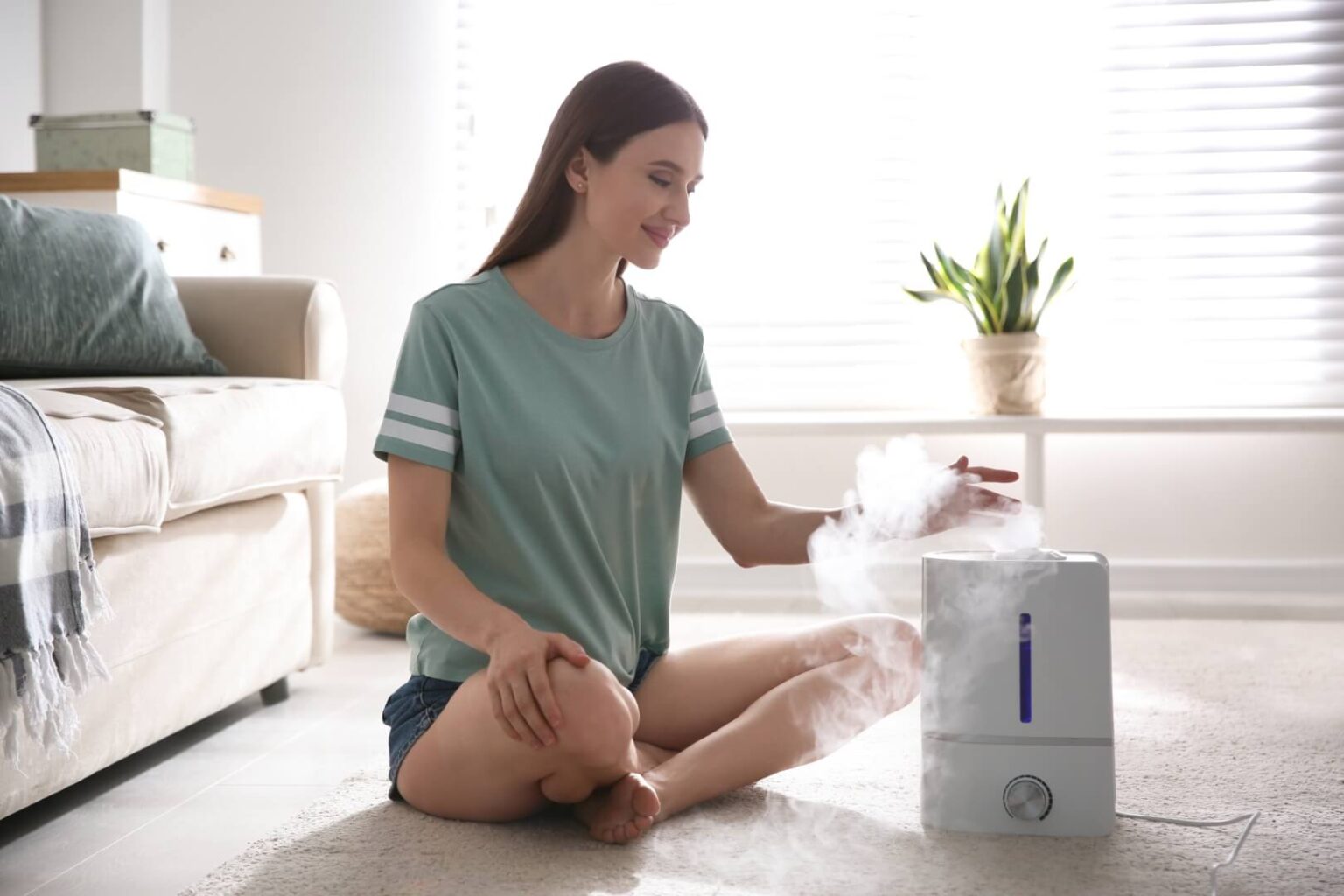 A woman sits cross-legged on the floor in a living room, placing her hand above the mist coming from a white humidifier. Sunlight streams through window blinds—an inviting scene for anyone considering humidifier installation in San Diego.