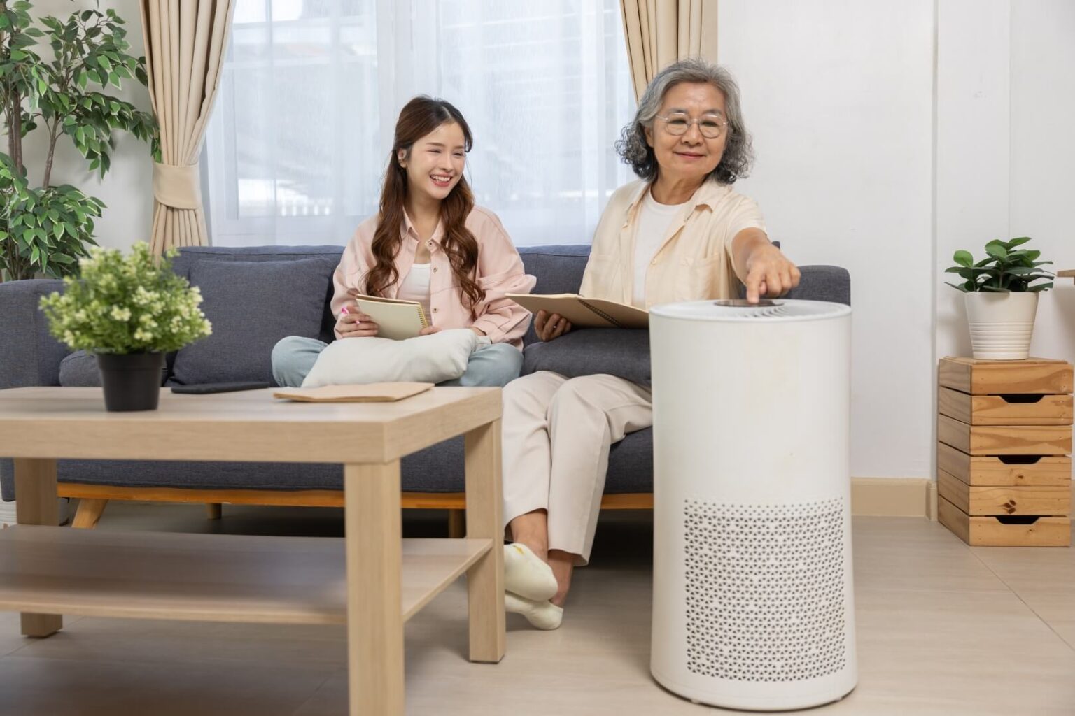 An older woman turns on a white air purifier while sitting on a couch with a younger woman, both holding tablets. They relax in a bright living room—highlighting the benefits of indoor air quality service San Diego residents trust.