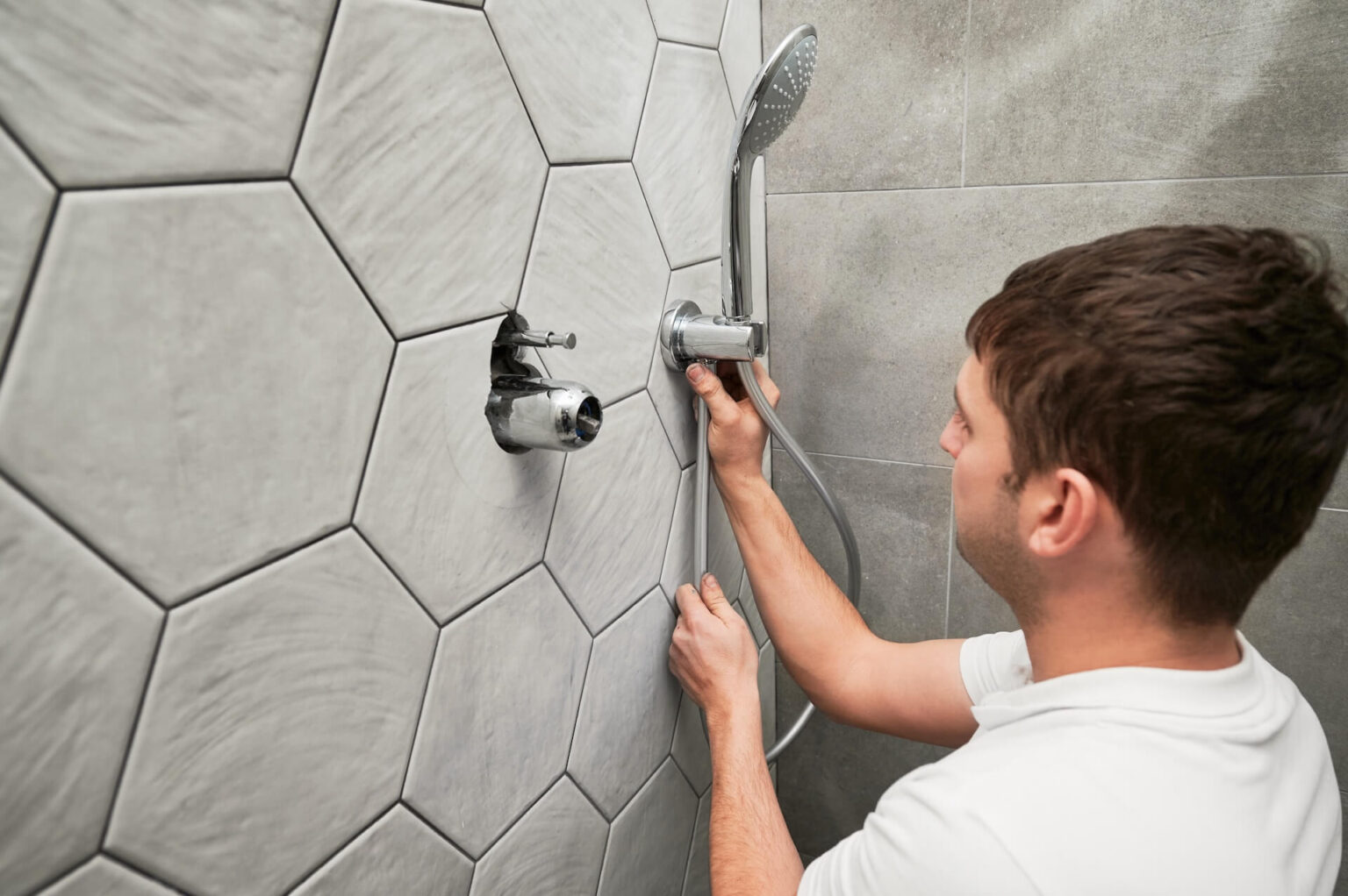 A man in a white shirt installs or repairs a handheld showerhead on a tiled bathroom wall with large, hexagonal gray tiles, showcasing the skill expected from expert Kitchen Plumbing Install San Diego professionals.