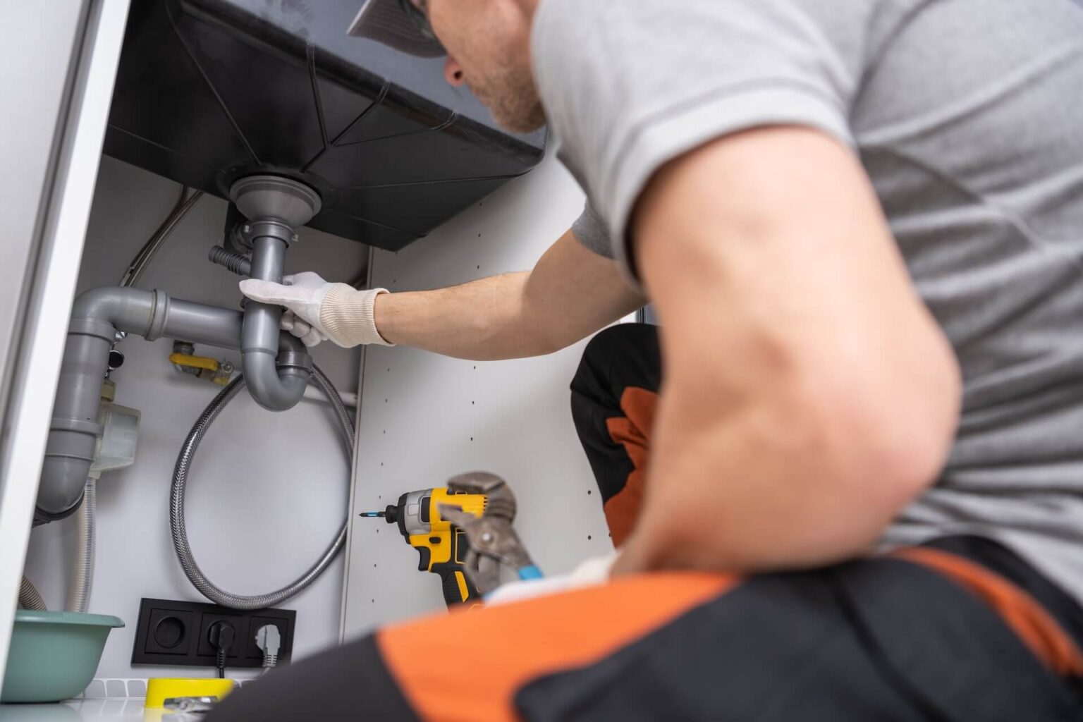 A person wearing gloves and work clothes uses tools to inspect or fix kitchen plumbing pipes under a sink. For expert Kitchen Plumbing Install San Diego services, a power drill and other equipment are ready inside the cabinet.