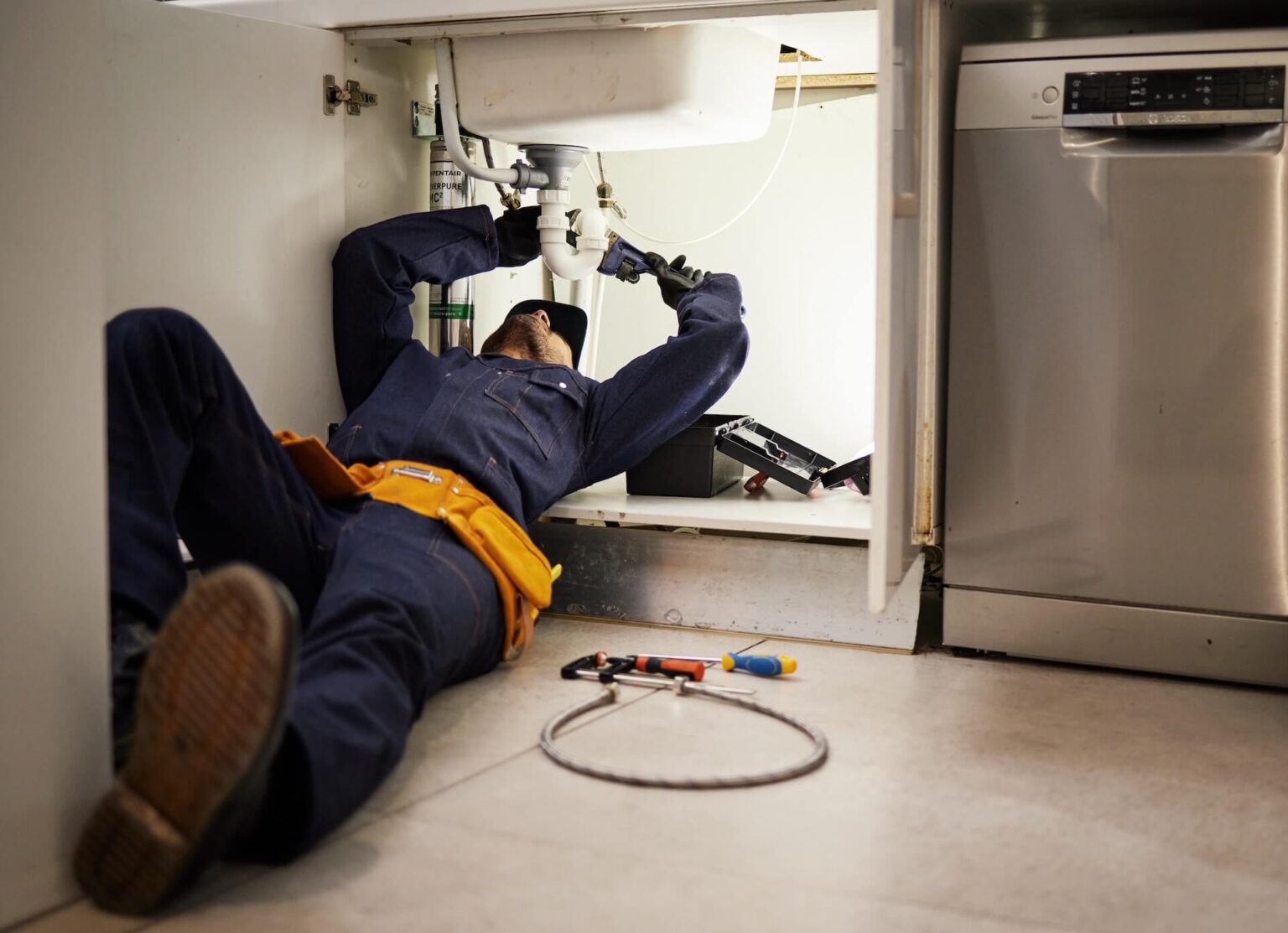 A plumber in a navy uniform lies on the floor fixing pipes under a kitchen sink. Tools, including pliers and a wrench, are spread out nearby. A dishwasher is installed next to the sink cabinet during this Kitchen Plumbing Repair San Diego.