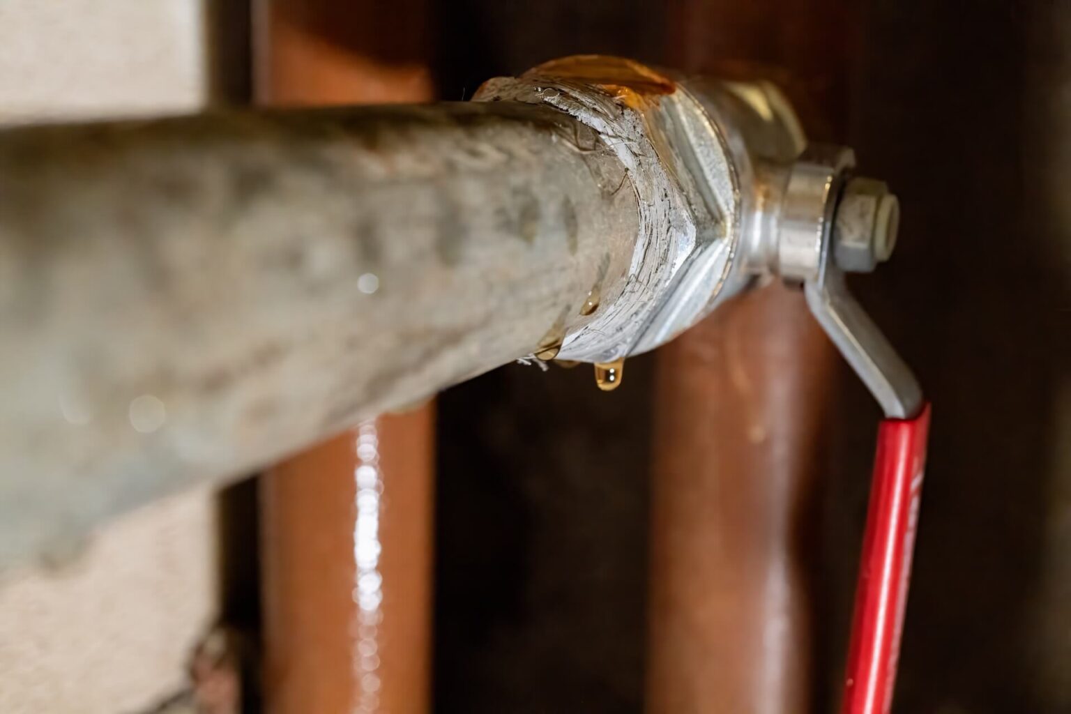 Close-up of a metal pipe with a valve handle and visible water leak, as shown by droplets forming around the connection—a scenario where Leak Detection San Diego services could help. Two brown pipes are blurred in the background.