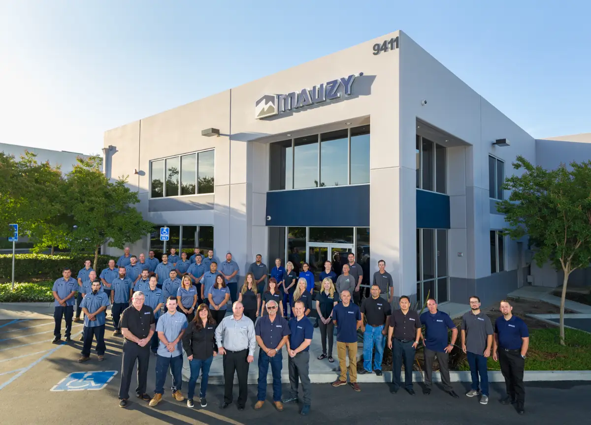 A group of people pose for a photo outside the modern Mauzy building, its name displayed above the entrance. On this sunny day, Sherlock joins men and women gathered in front of the white and gray structure.