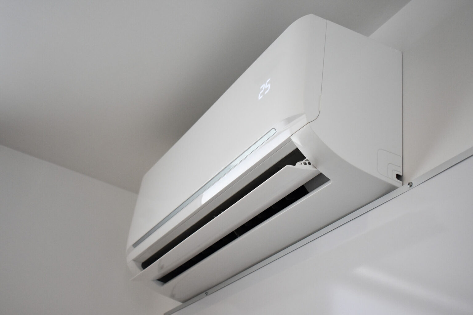A wall-mounted white air conditioner, from a mini split install San Diego service, is turned on and displays 25 on its digital screen. The vents are open, and the unit is installed near the ceiling in a plain, white room.